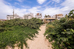 Uma trilha de areia conduz a um edifício com vários andares e varandas, cercado por vegetação densa.