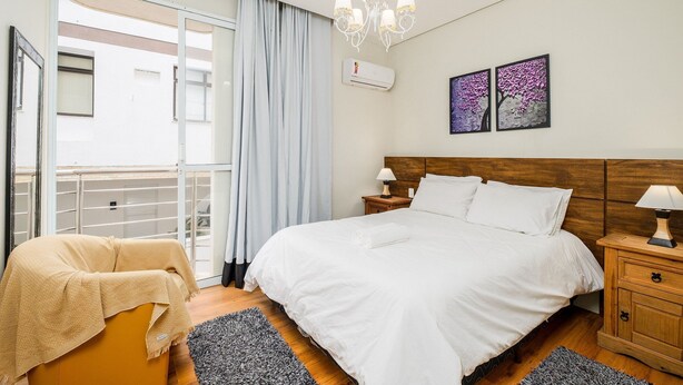 A bedroom with a bed, a nightstand, a lamp, a ceiling light, a window with curtains, and artwork on the wall. - Apart Foot on Sand for Couples in Floripa Ida105 (Florianópolis)