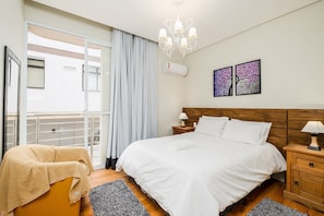 A bedroom with a large bed, a wooden nightstand, a chandelier, and a balcony with a sliding door.