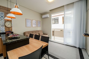 A modern living room with a sofa, dining table, and chairs. There is a kitchen area with a stove and a clock. The room has a large window with curtains and an air conditioning unit on the wall.