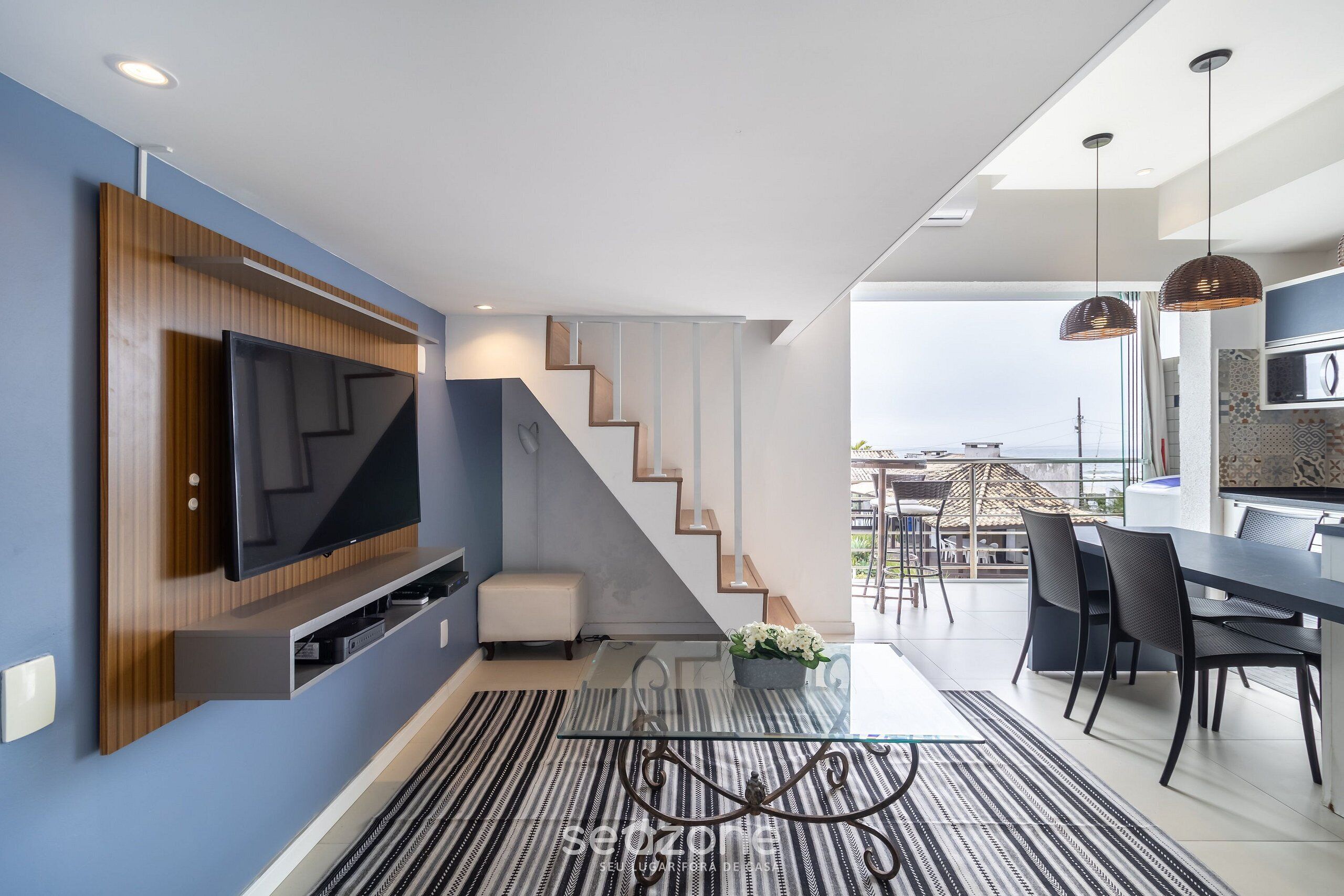 A modern living room with a flat-screen TV, a staircase, a dining area, and a view of the city.