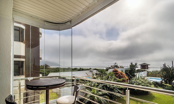 A balcony with a table and chairs, overlooking a swimming pool and lush greenery.