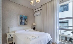 A hotel room with a large bed, a nightstand, an air conditioning unit, a window with a view of a building, and a painting on the wall. - Excellent Beachfront apt With Barbecue Ida101 (Florianópolis)