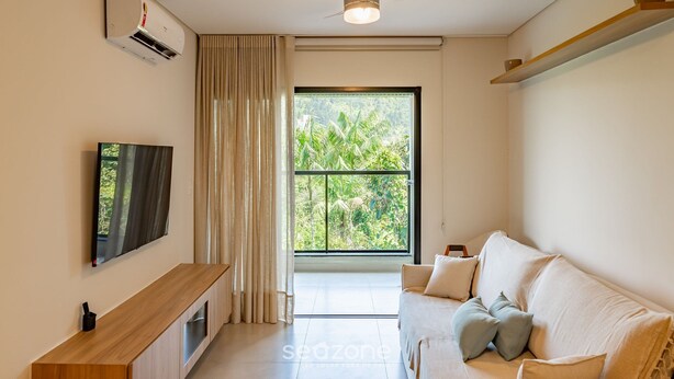 A modern living room with a flat-screen TV, a ceiling fan, a large window with a view of greenery, and a sofa with cushions. - Apartment w/ Barbecue 290m From the Beach Hre0035 (Ubatuba)