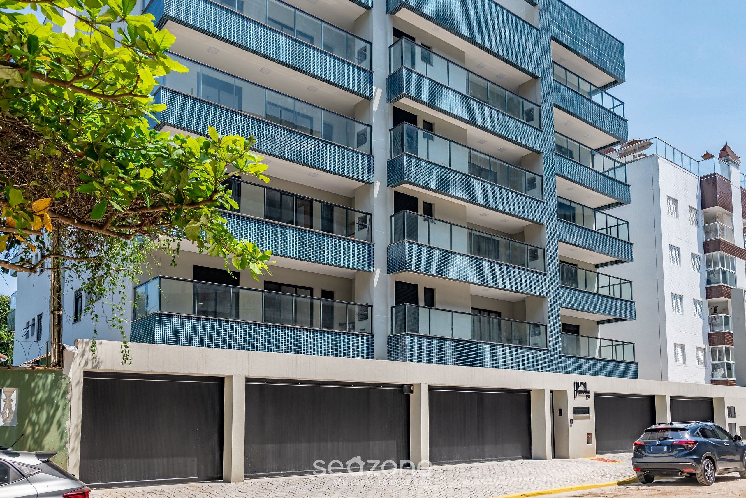 Un edificio residencial de varios pisos con balcones, un área de garaje y un árbol en primer plano.