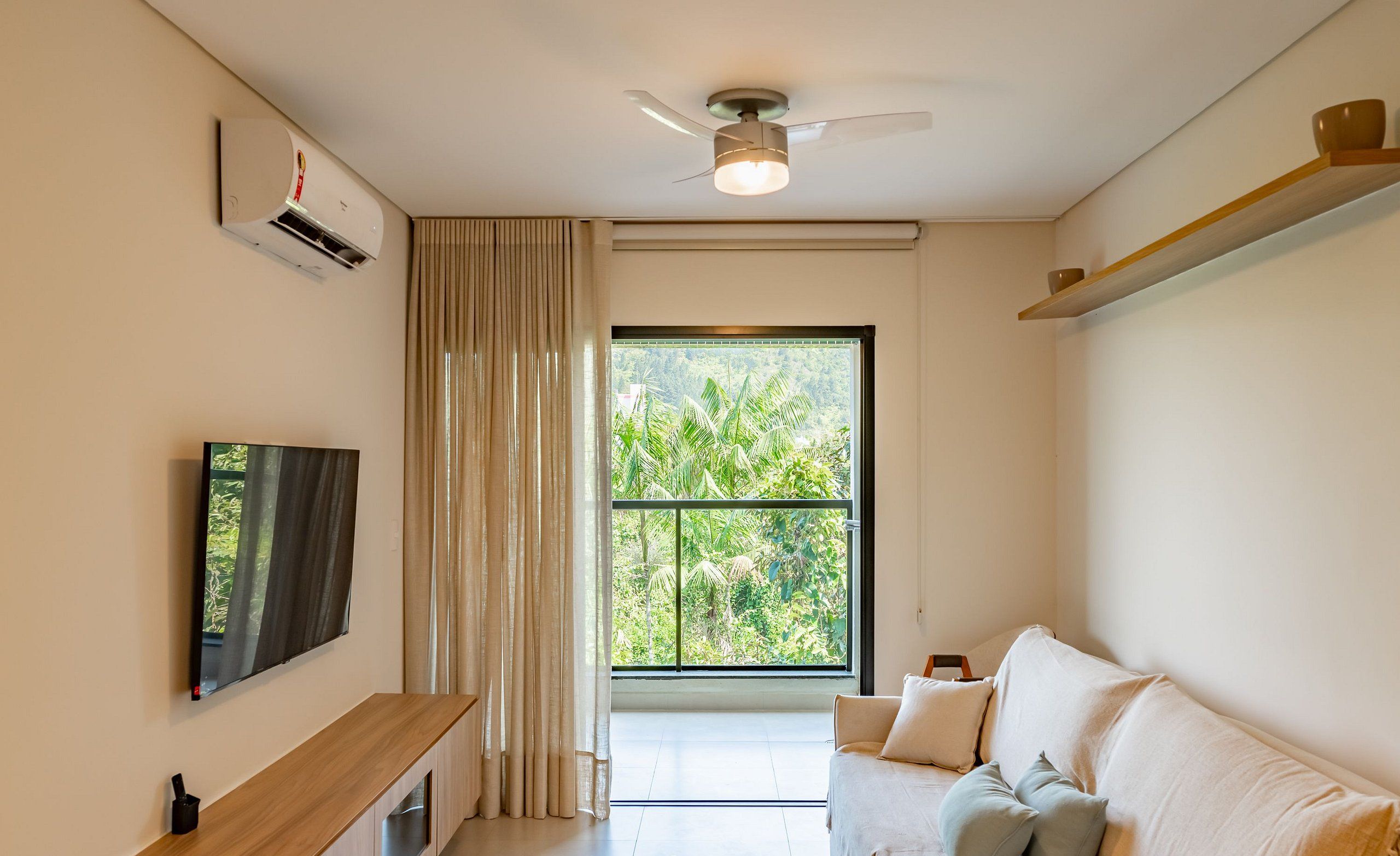 A modern living room with a flat-screen TV, a ceiling fan, a large window with a view of greenery, and a sofa with cushions.
