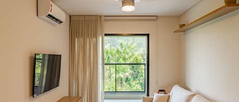 A modern living room with a flat-screen TV, a ceiling fan, a large window with a view of greenery, and a sofa with cushions.
