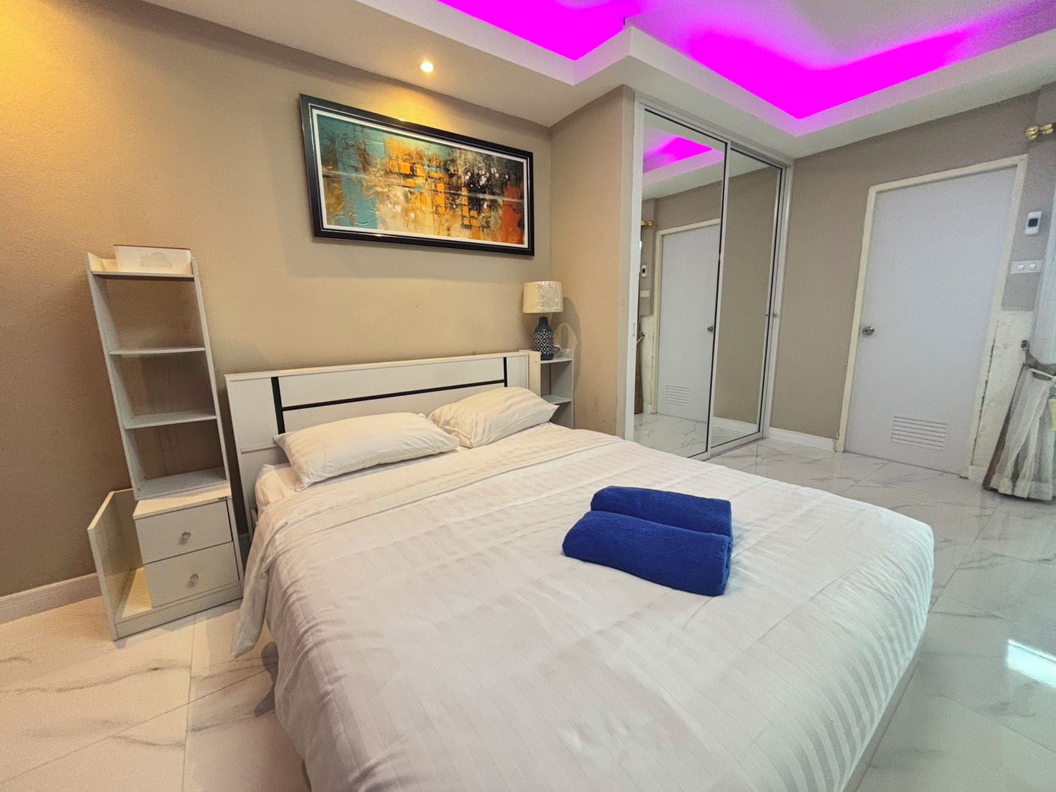 A hotel room with a bed, a nightstand, a shelf, a painting, and a door.