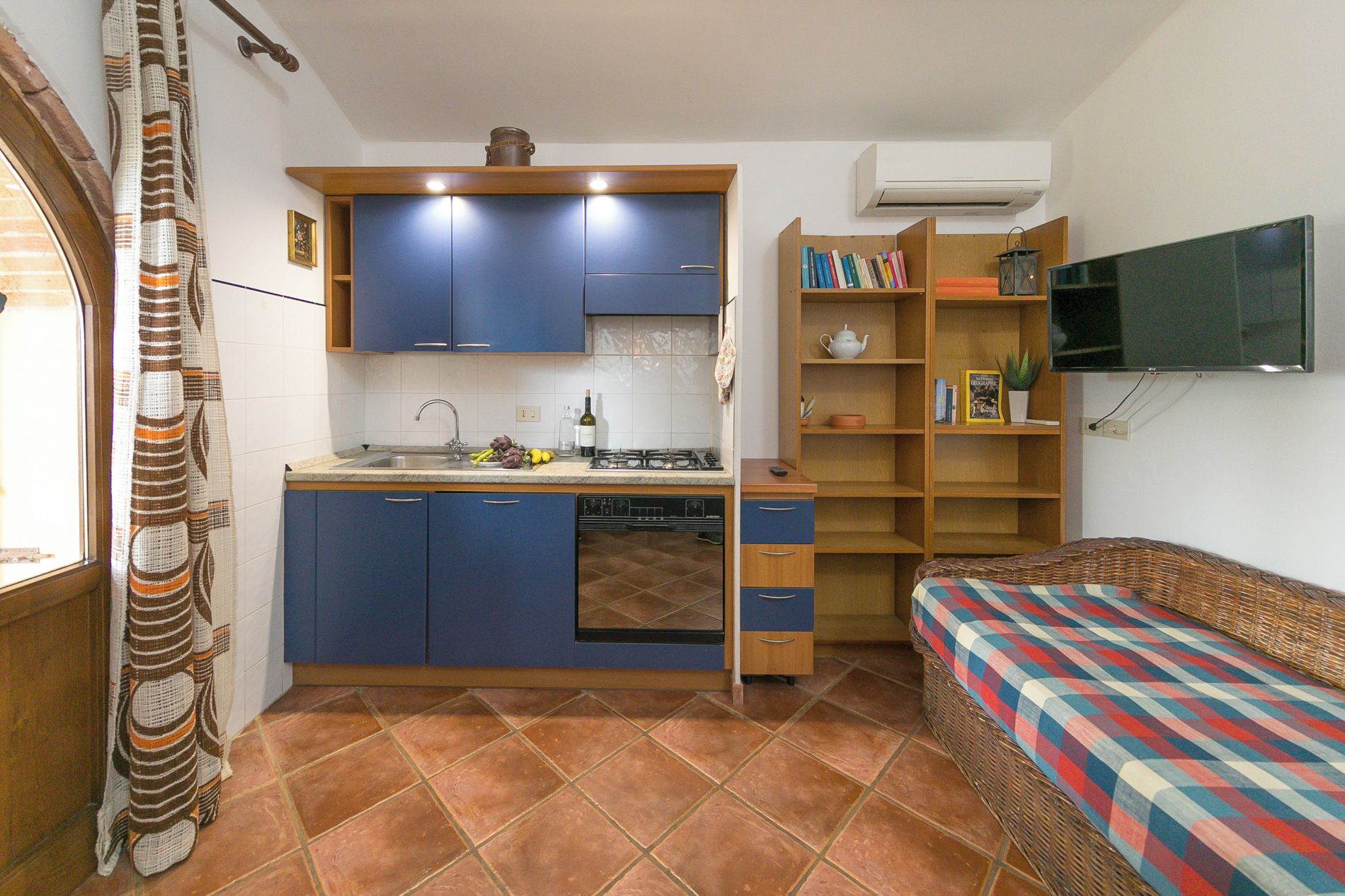 A compact kitchen with blue cabinets, a gas stove, and a sink. There is a wicker sofa with a patterned cushion and a television mounted on the wall.