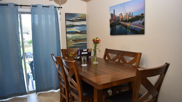 A dining area with a wooden table, four chairs, a vase with flowers, a painting on the wall, and a view of a cityscape through a window. - Cozy Newly Renovated 3 Bedroom Home 5 min. to SoCo & 15 min. to Zilker Park (Austin)