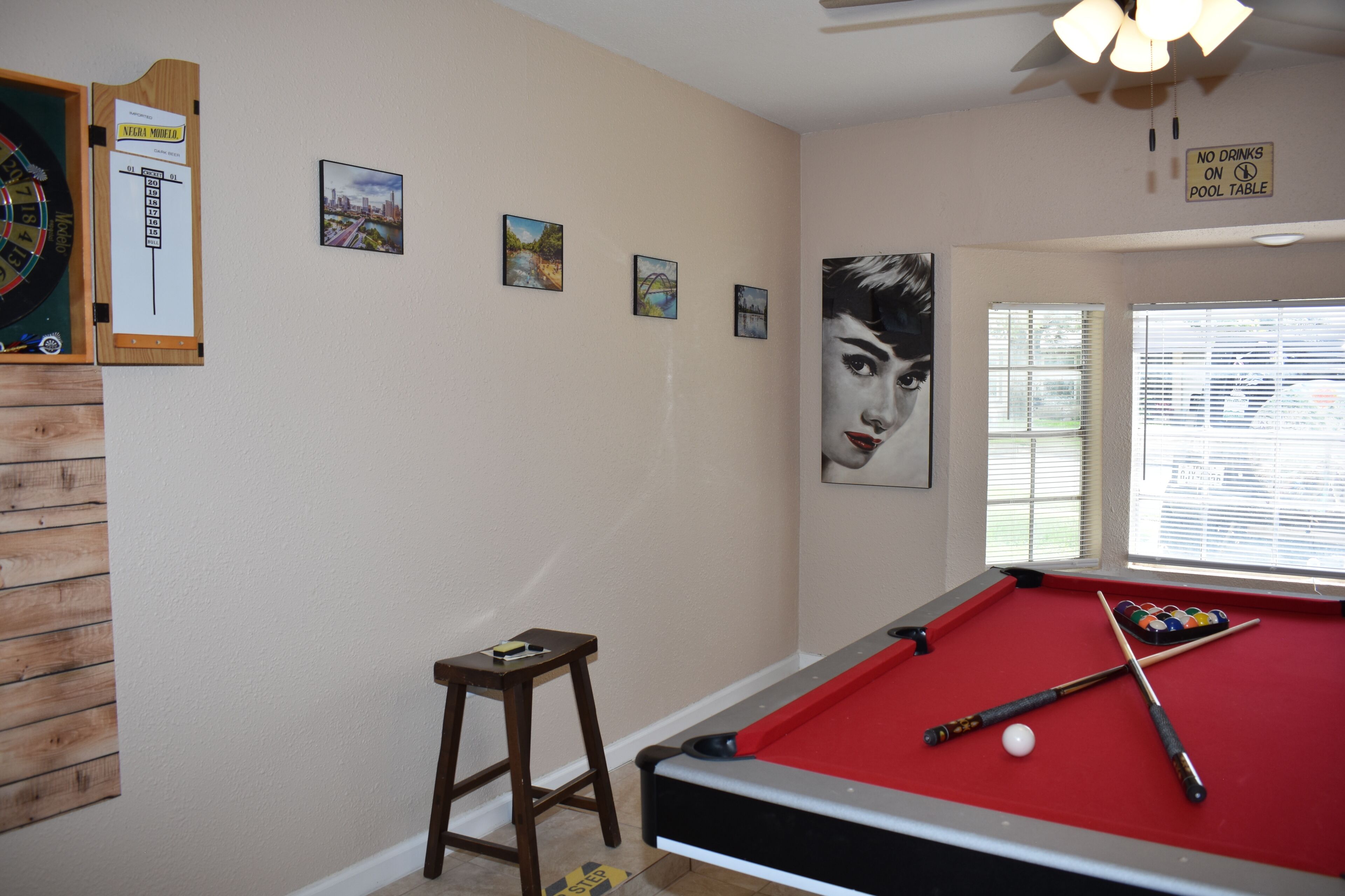 Une table de billard recouverte d’un drap rouge, un queue de billard et une bille. Il y a quatre tableaux encadrés au mur, un jeu de fléchettes et un panneau indiquant qu’il est interdit de poser des boissons sur la table de billard.