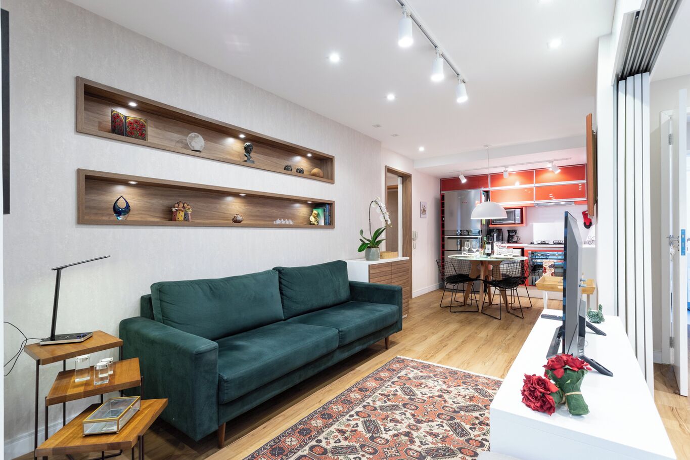 Una sala de estar moderna con un sofá verde, estanterías de madera con objetos decorativos, una zona de comedor con mesa y sillas, y una cocina al fondo.