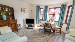 A living room with a TV, a dining table with chairs, a sofa, and a wooden cabinet with decorative items. - Flat 9 (Swanage)