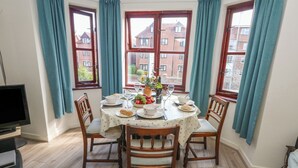 A dining area with a round table set for four, surrounded by wooden chairs. There is a television on the left side of the room. - Flat 9 (Swanage)