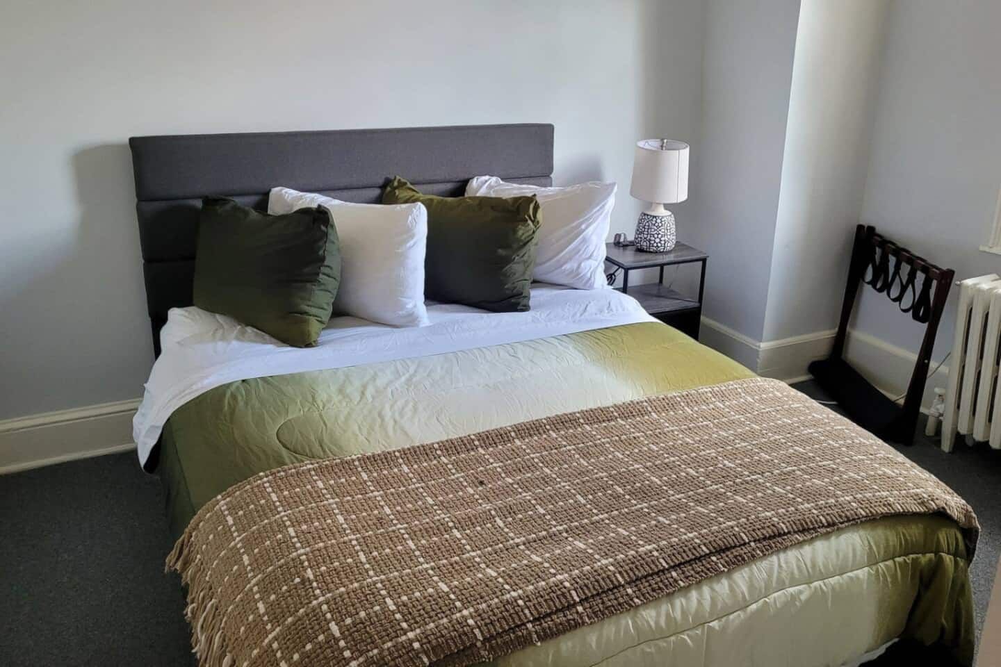Una cama bien hecha con un cabecero gris, almohadas blancas y verdes, una manta marrón y una mesita de noche con una lámpara.