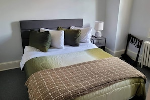 Een net opgemaakt bed met een grijze hoofdbord, witte en groene kussens, een bruine deken en een nachtkastje met een lamp.