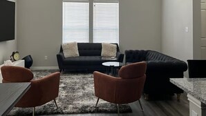 Ett modernt vardagsrum med en svart hörnsoffa, två fåtöljer, ett soffbord och en takfläkt. Det finns stora fönster med persienner, en platt-TV som är monterad på väggen och en köksdel med en bänkskiva i marmor.