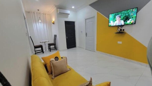 Un salon avec un canapé jaune, un téléviseur à écran plat fixé au mur et un coin repas comprenant des chaises et une table.