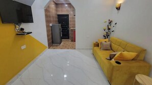 Un salon moderne avec un canapé jaune, un téléviseur à écran plat fixé au mur et un coin cuisine visible à travers une arche.