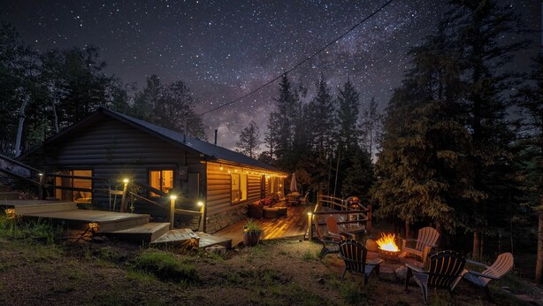 Una cabaña de troncos con terraza, mobiliario exterior y un pozo de fuego.