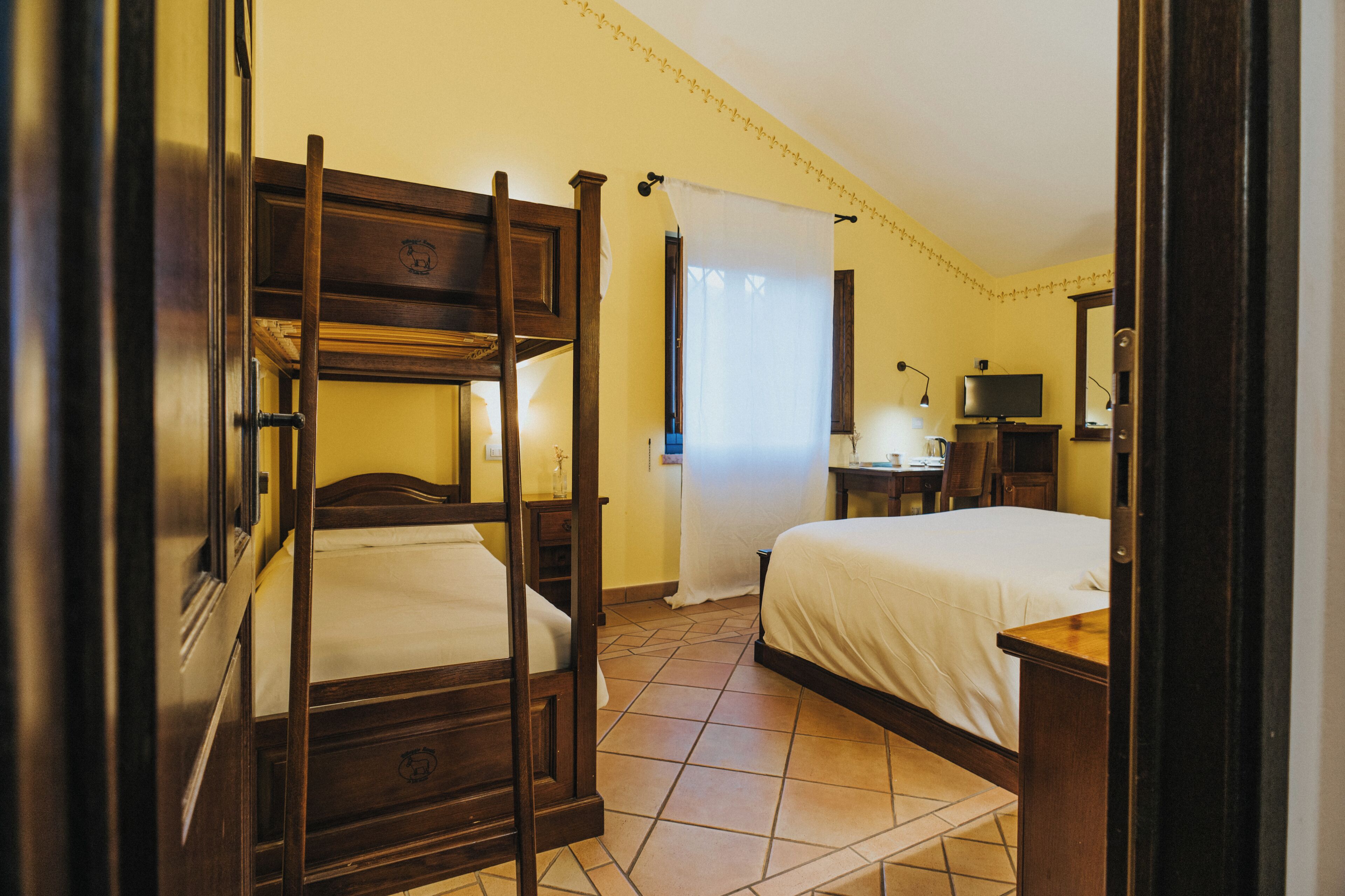 Una habitació amb un llit superposat, un escriptori amb cadira, una televisió i una finestra amb cortines.