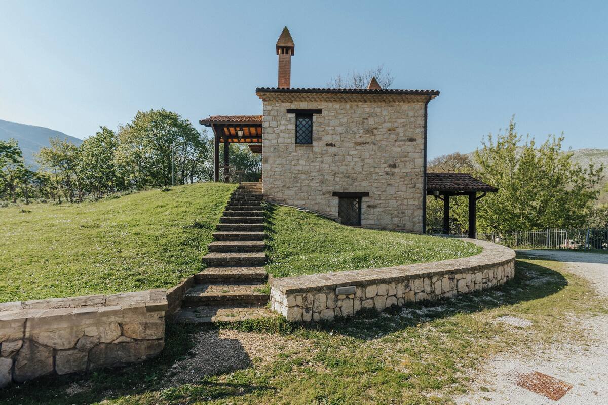 Ein Steinhaus mit einem Turm, einer überdachten Holzkonstruktion und einer Steintreppe, die einen grasbewachsenen Hügel hinaufführt.