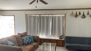 A living room with a brown sofa, a glass coffee table, and a dark grey sofa. There is a window with curtains and a ceiling fan.