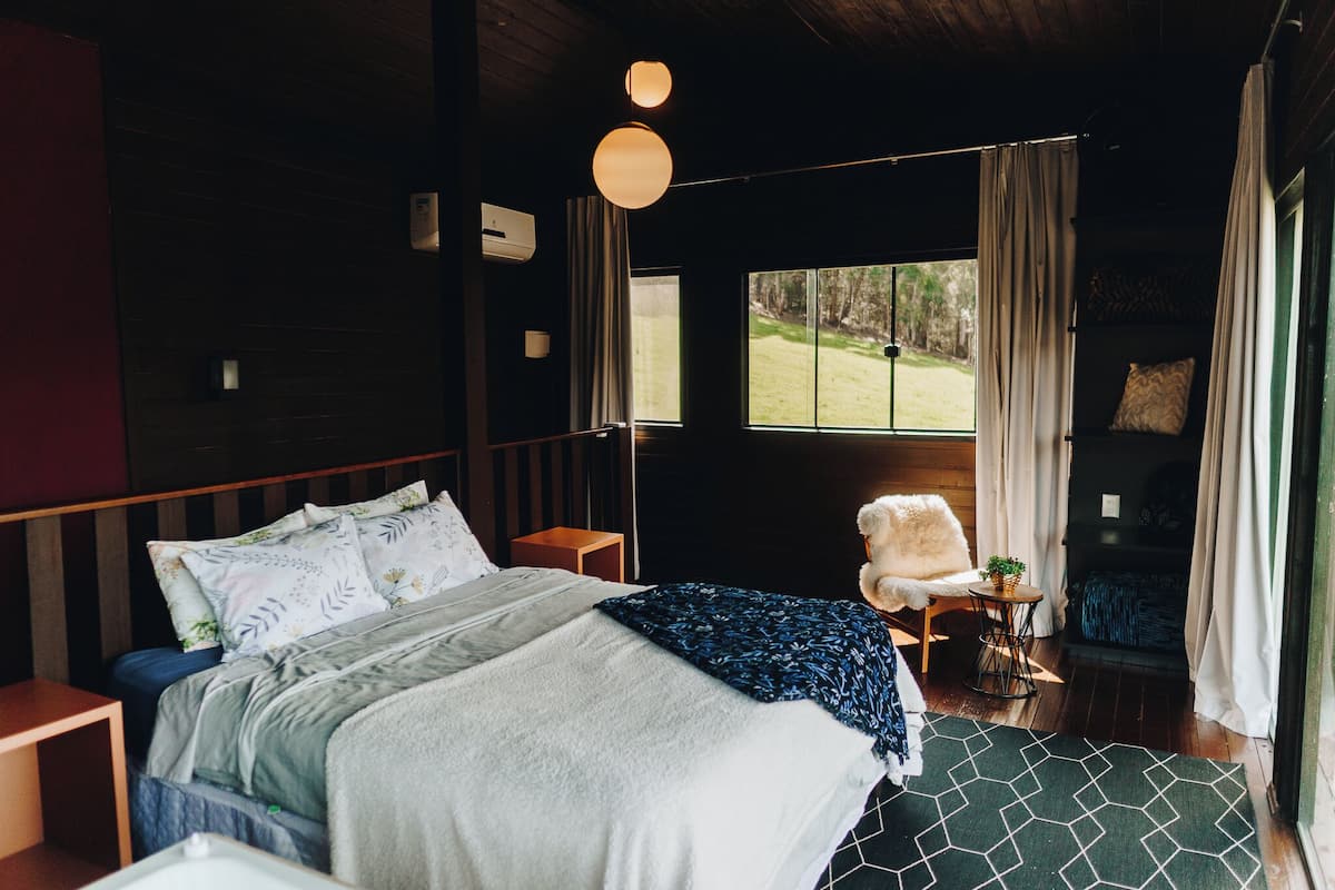 Une chambre à coucher avec un grand lit, une petite table sur laquelle est posée une plante, et une vue sur l’extérieur par une fenêtre.
