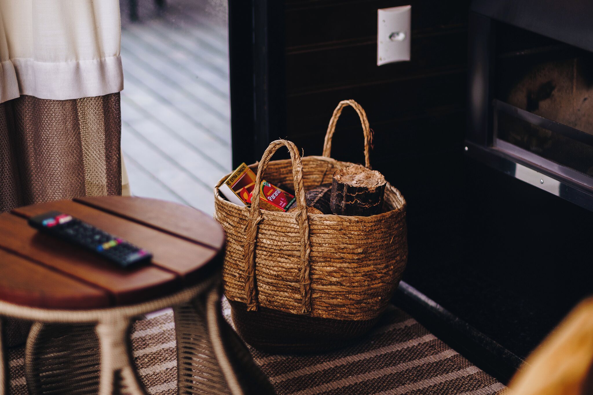 Una cesta de mimbre con aperitivos y un control remoto sobre una mesa de madera junto a una ventana.