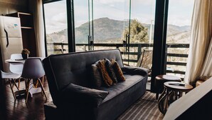 A modern living room with a dark grey sofa, white chairs, and a small table with a plant. Large windows offer a view of a mountainous landscape. - Loft Felicidade  (Urubici)
