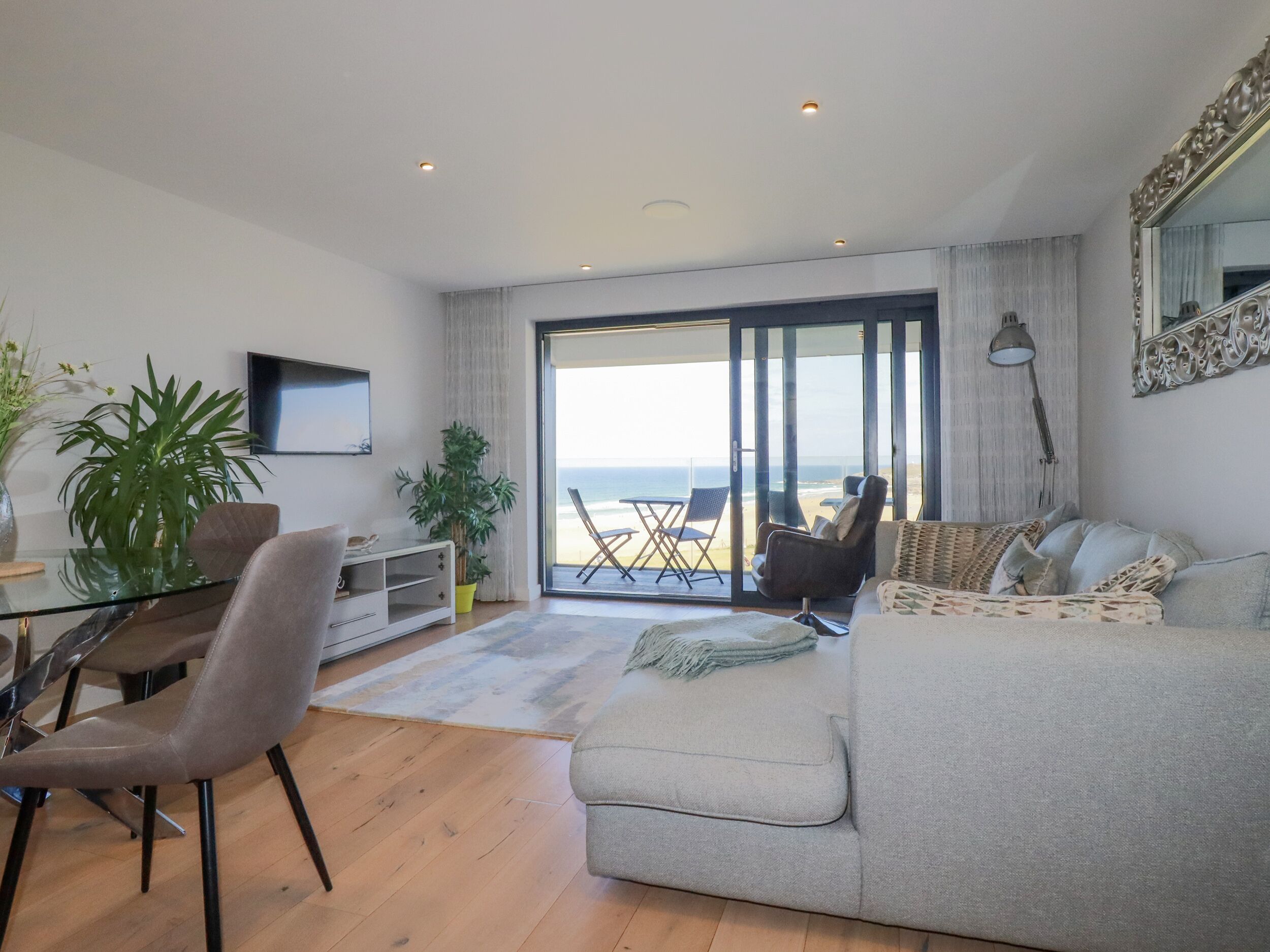 A modern living room with a sofa, a dining table, and chairs. There is a large window with a view of the beach.