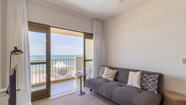A modern living room with a grey sofa, two pillows, a small side table, and a TV. A large sliding glass door leads to a balcony with a view of the sea and a white tent. - Great Beachfront Apartment Lbm306 (Florianópolis)