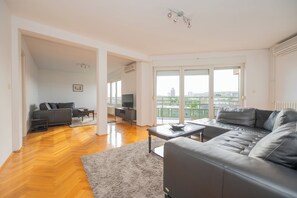 En rummelig opholdsstue med sofa, sofabord og fjernsyn. Der er en balkon med udsigt over byen.