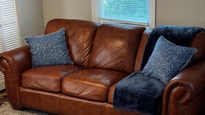 A brown leather sofa with two patterned pillows and a dark throw blanket, placed on a patterned rug in a room with a window and blinds. - The Porter Blue (Augusta)