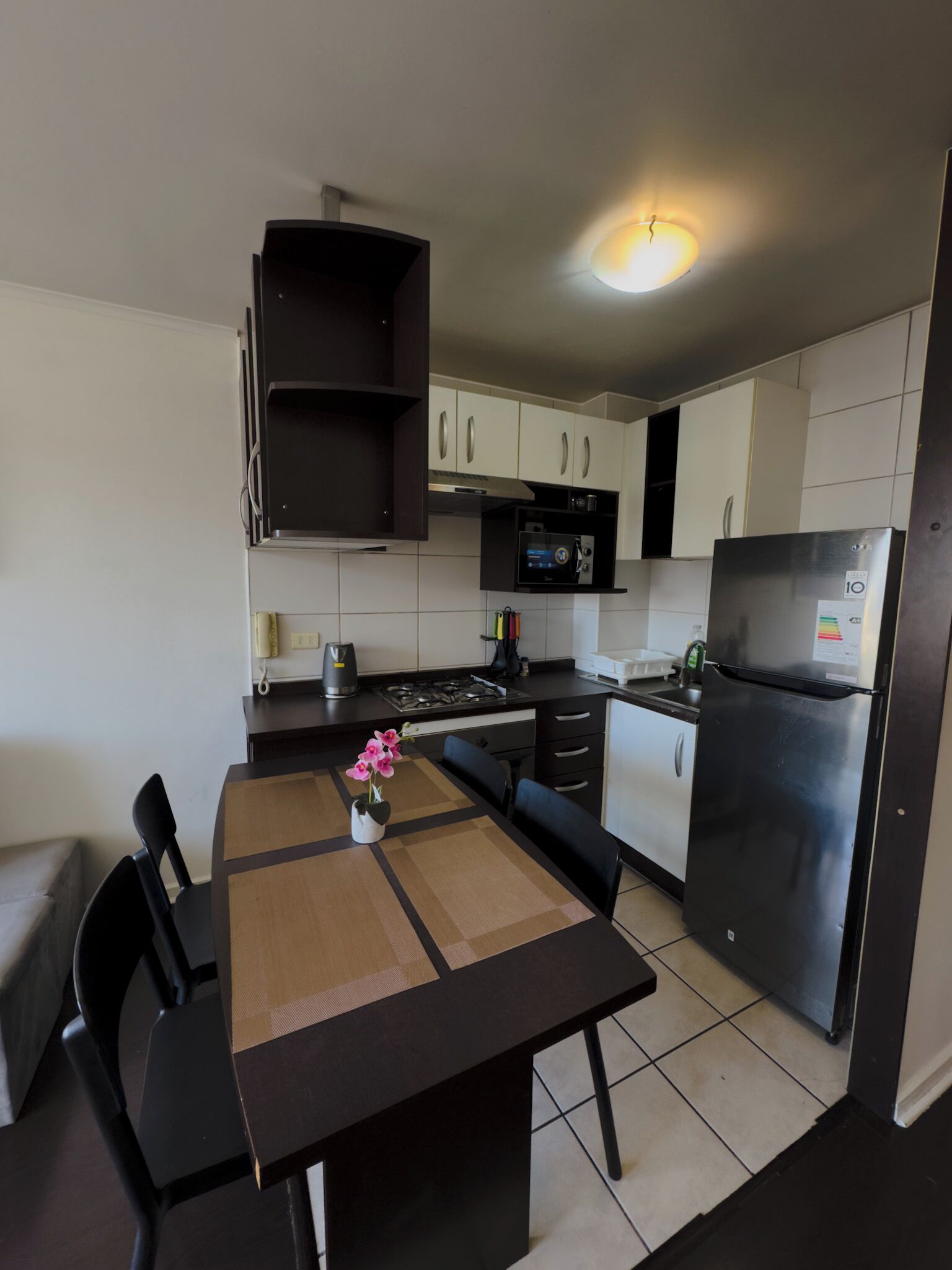 Una cocina moderna con armarios blancos, un refrigerador negro y un microondas. Hay una mesa de comedor con sillas y un florero con flores.