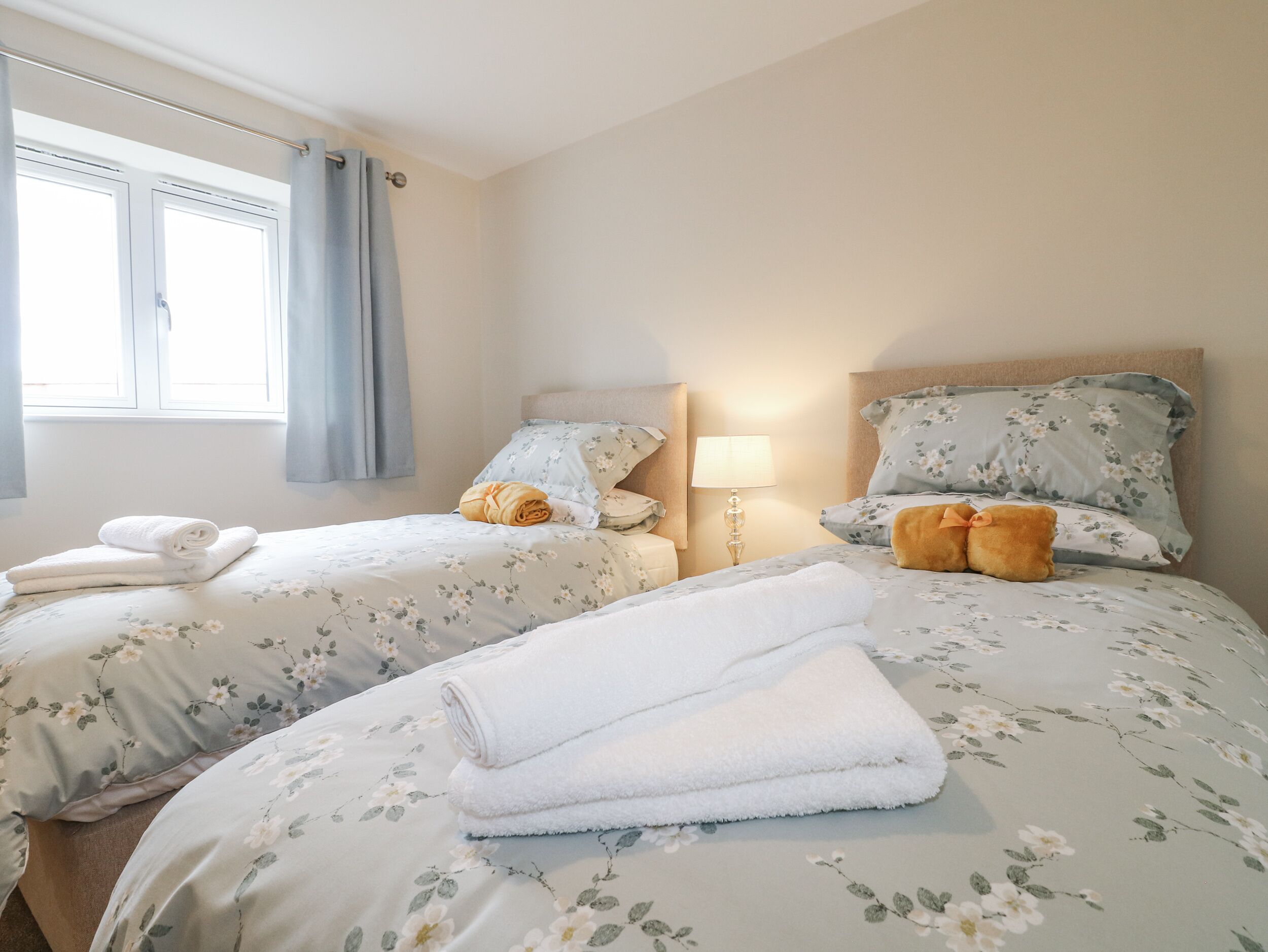 A hotel room with two beds, each with floral bedding and pillows, a bedside table with a lamp, and a window with curtains.