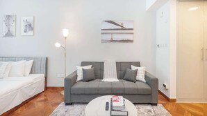 A modern living room with a grey sofa, a white coffee table, and a floor lamp. There are framed pictures on the wall and a door to the right. - Production City Sleeps 4 With Water Views (Dubai)