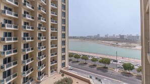 A high-rise building with multiple balconies overlooking a coastal area with a road and parked cars. - Production City Sleeps 4 With Water Views (Dubai)