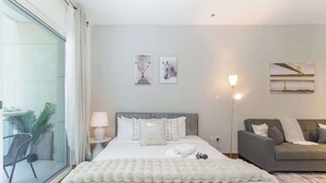 A modern bedroom with a large bed, a grey sofa, and a balcony with plants. - Production City Sleeps 4 With Water Views (Dubai)