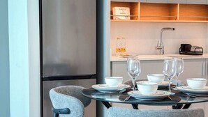 Eine kompakte Küche mit einem modernen, edelstahlfarbenen Kühlschrank, einem eingebauten Backofen und einer Mikrowelle. Es gibt eine kleine Essecke mit zwei Stühlen und einem Tisch für zwei Personen, komplett mit weißen Tellern, Schüsseln und Weingläsern.