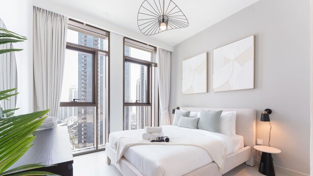 Ein modernes Schlafzimmer mit einem großen Bett, einem Nachttisch, einer Lampe und Blick auf die Stadt durch große Fenster.