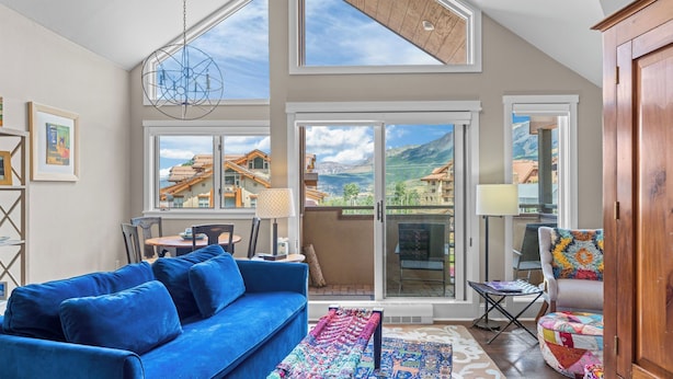 A modern living room with a blue sofa, a colorful rug, and a view of mountain houses through large windows. - Blue Mesa Lodge 40A by Avantstay Ski In/ Ski Out Unit w/ Balcony! (Telluride)