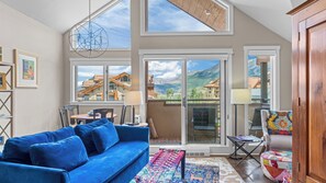 Uma sala de estar moderna com um sofá azul, um tapete colorido e vista para casas de montanha através de grandes janelas.