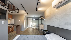 Un dormitorio moderno y funcional con cama, mesitas de noche y un armario. Se ve una pequeña cocina con estufa y armarios al fondo.