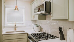 A kitchen with white cabinets, a gas stove, a microwave, and a window with blinds. - 2BR/1BA in Golf Cart District near Beaches and Downtown! The Beachcomber A (Ocean Springs)