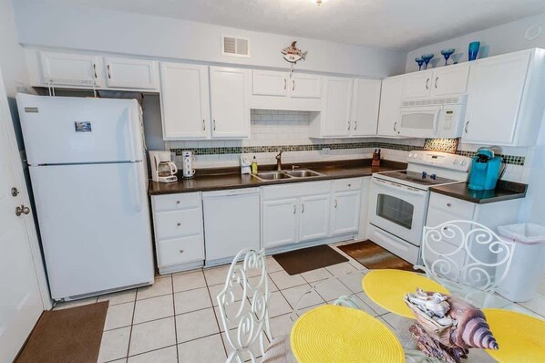 A kitchen with white cabinets, a refrigerator, a microwave, a stove, and a sink. There is a dining table with yellow placemats and a decorative shell.