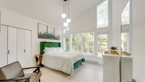 Een moderne slaapkamer met een groot bed, een groen hoofdeinde en uitzicht op bomen door grote ramen.