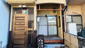 Een woongebouw met een houten voordeur, een klein portaal met betegelde vloer en een balkon met metalen hekwerk.
