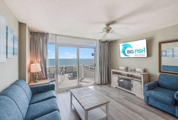 A spacious living room with a large sliding glass door leading to a balcony, a flat-screen TV mounted on the wall, and a ceiling fan.