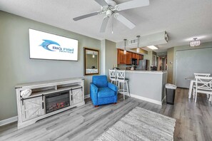A modern living room with a fireplace, a blue armchair, a dining area with white chairs, and a kitchen in the background.